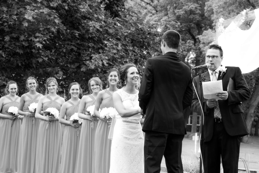 Young couple on wedding day, bride and bridesmaids smiling at groom at the alter with pastor officiating