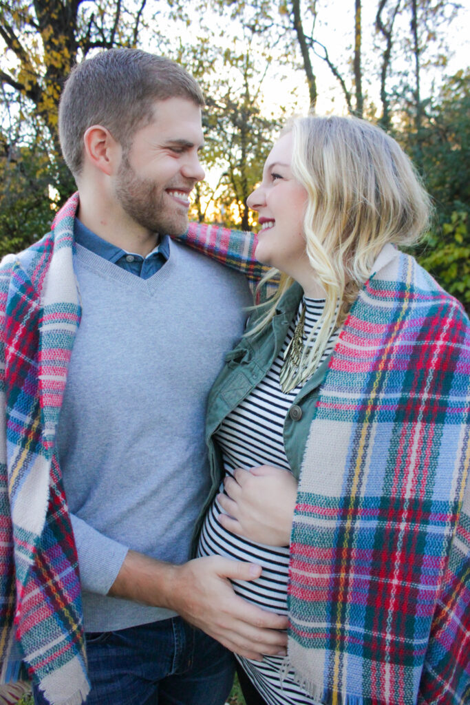 Couple wrapped in a plaid scarf looking at each other smiling, husband's hand is on wife's pregnant belly