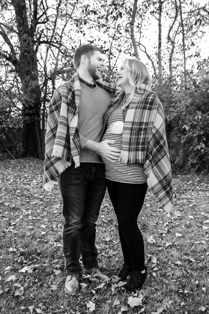 Couple wrapped in a plaid scarf looking at each other laughing, husband's hand is on wife's pregnant belly