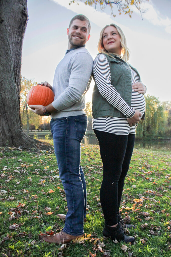 Couple back-to-back looking over their shoulders at each other, wife is holding belly and husband is holding pumpkin