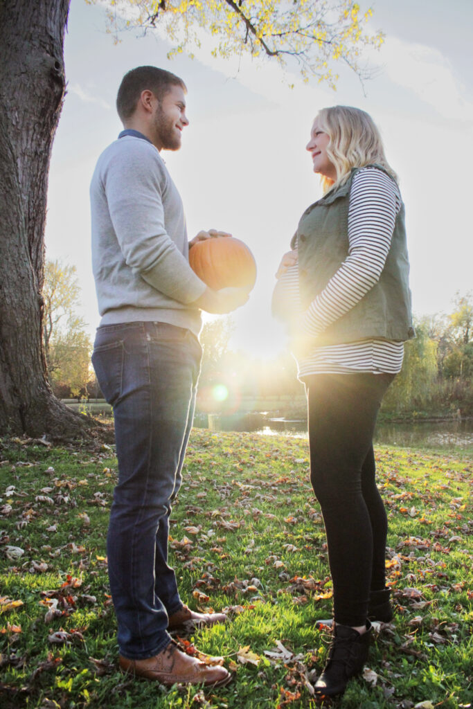 Couple back-to-back looking over their shoulders at each other, wife is holding belly and husband is holding pumpkin