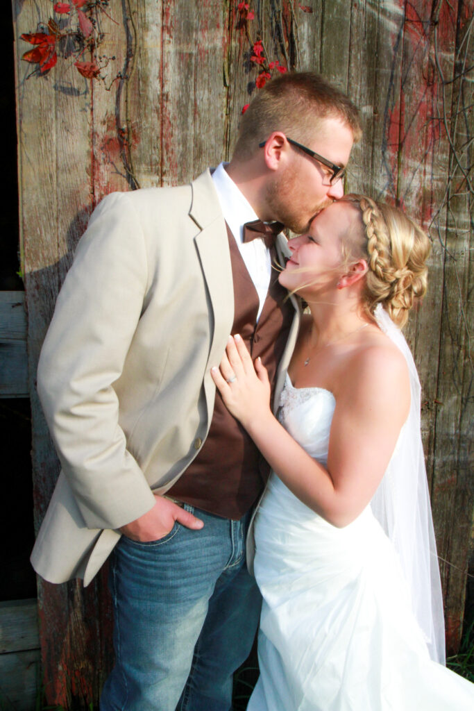 Groom kisses forehead of bride with rustic barn as background