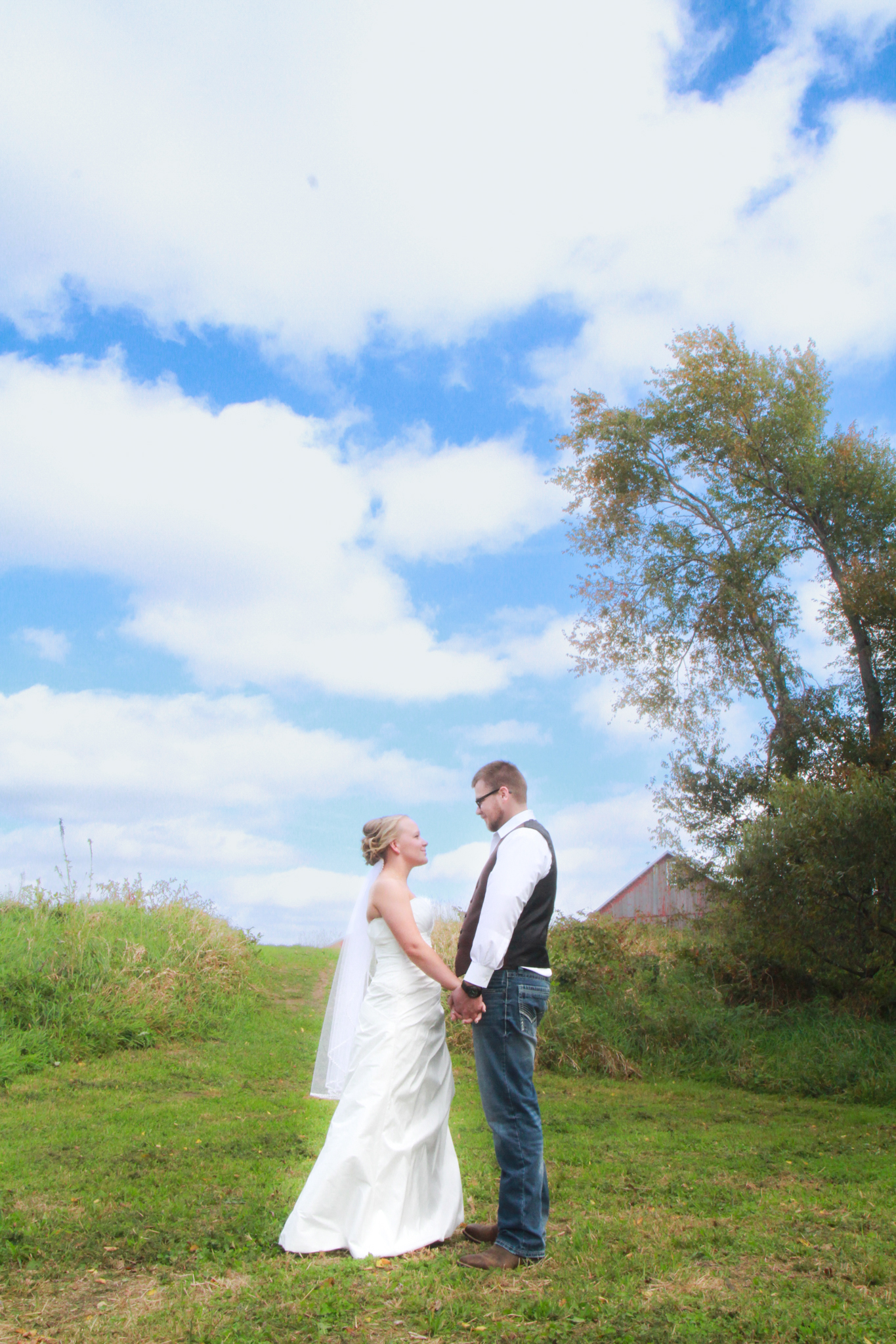 Bride and groom hold hands and look into each other's eyes with big blue sky and rustic barn in the background