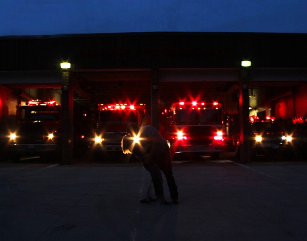 Man dips woman for kiss in front of lights of firetrucks, backlight