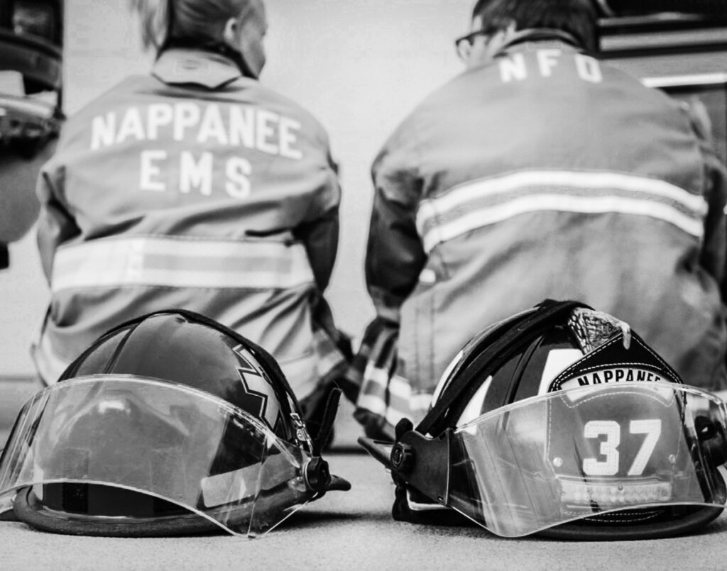 Couple sits facing away from camera, woman is wearing EMS jacket and man is wearing fire department jacket; individual associated helmets are in foreground