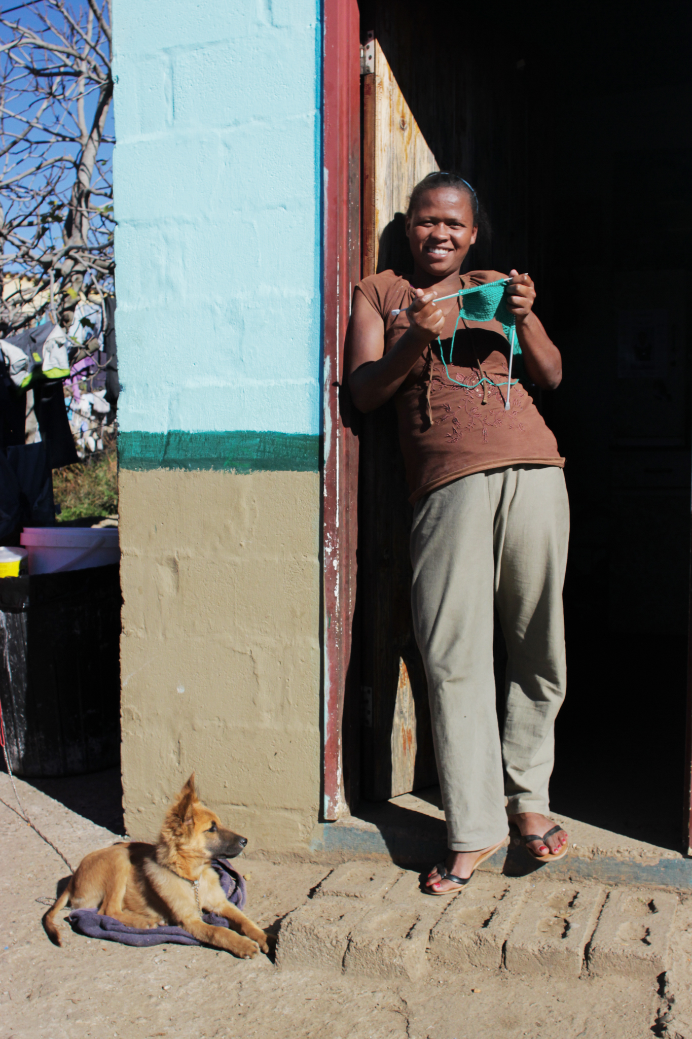 Woman from Blanco township in Goerge, South Africa standing barefoot and smiling in a doorway with her knitting, a small puppy laying at her feet