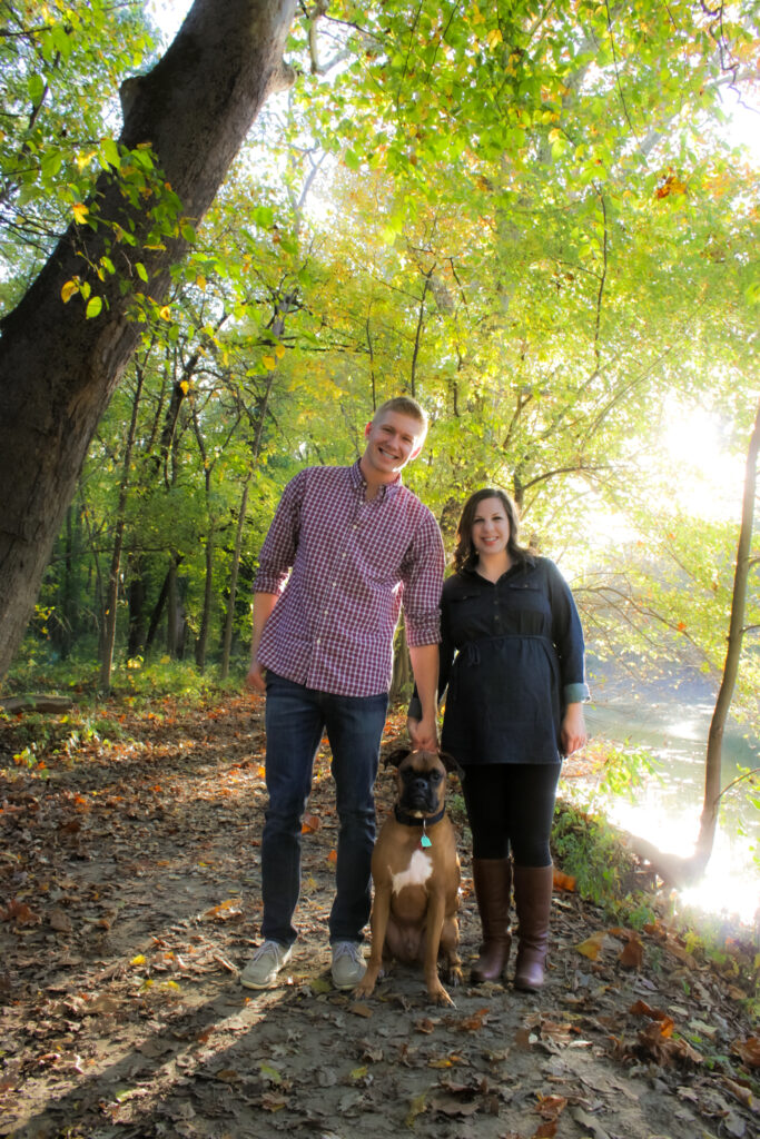 couple in maternity photo pose smiling with their boxer dog in the woods on a leafy path