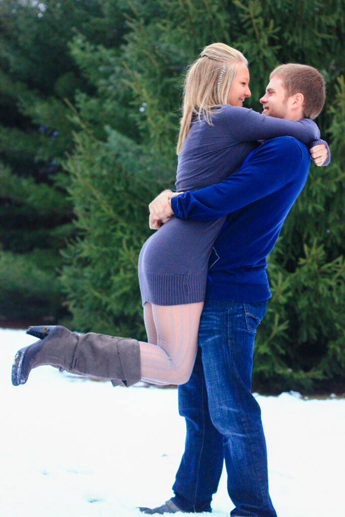 Young man holds up young woman as they smile at each other standing in the snow in front of pine trees