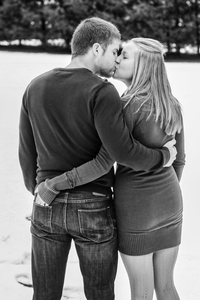 Young man and young woman facing away from camera with their arms around each other kissing while standing in snow in front of pine trees