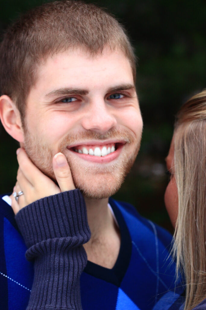 Young man smiles with young woman's hand with engagement ring holding his cheek