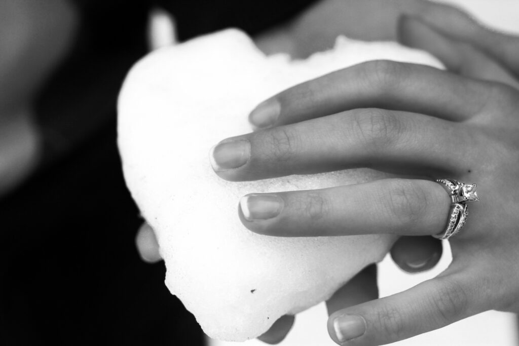 Young man and woman hold snow packed into the shape of a heart, woman's hand with engagement ring on top of heart