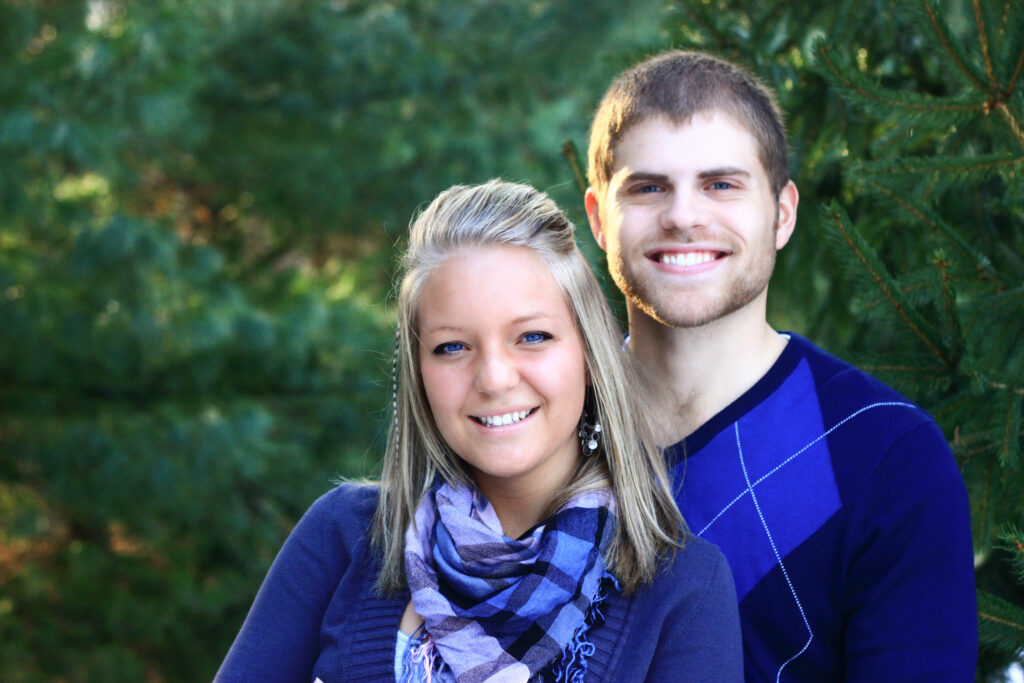 Young man and young woman smile at camera standing in front of pine trees