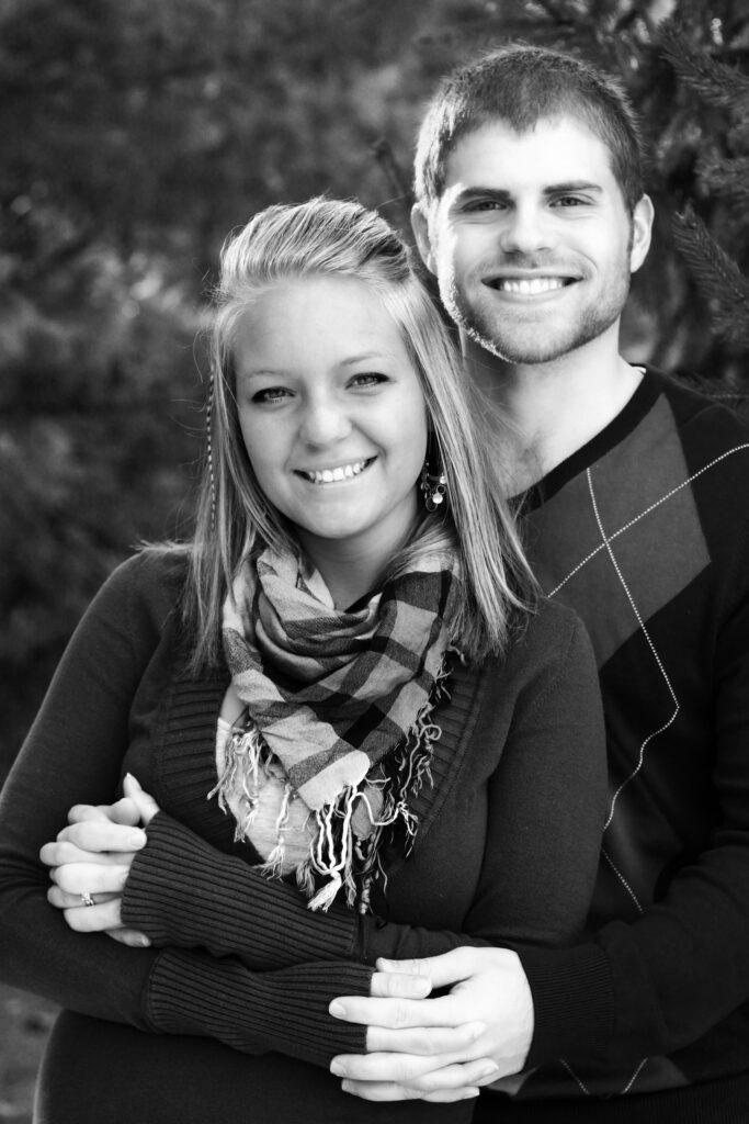 Young man and young woman smile at camera standing in front of pine trees