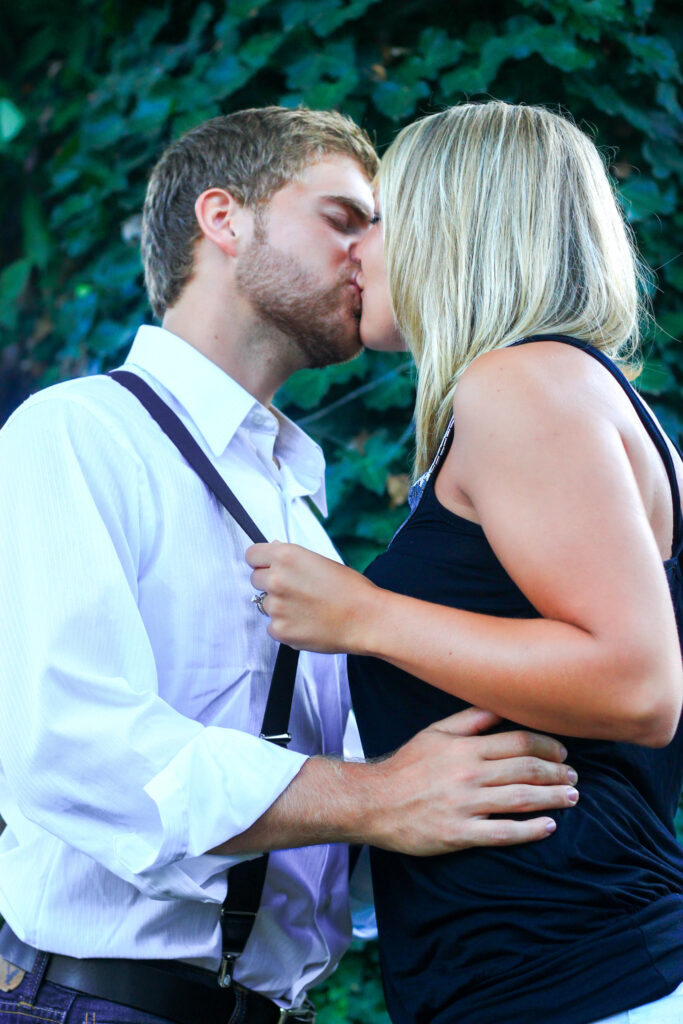 Young couple kissing in front of vine-covered tree, she is pulling him closer with suspenders