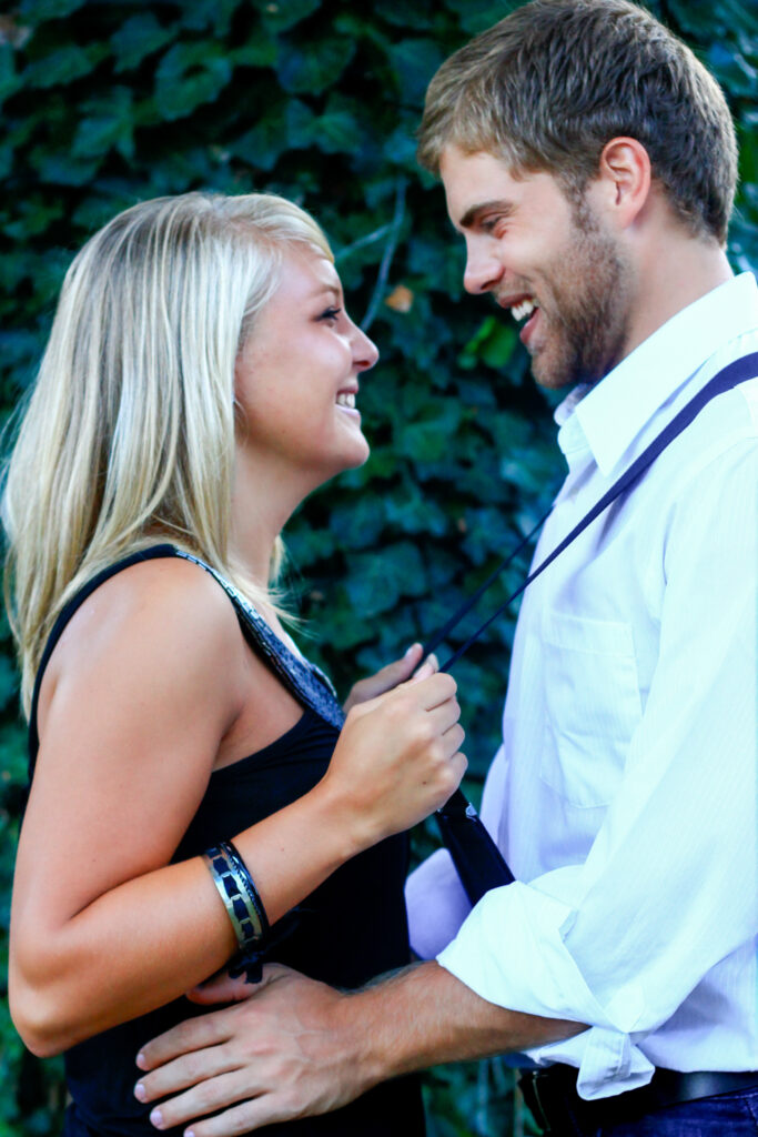 Young couple smiling at each other in front of vine-covered tree, she is pulling him closer with suspenders