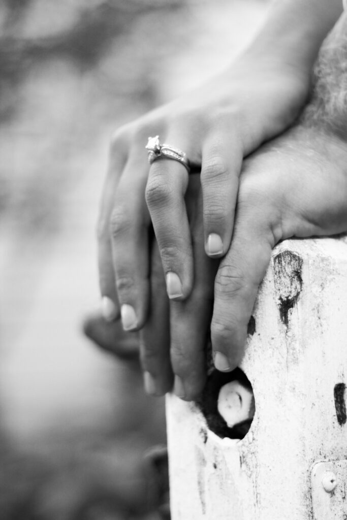 Hands of engaged couple are stacked on top of each other on a rustic fence pole, her hand on top with shiny engagement ring