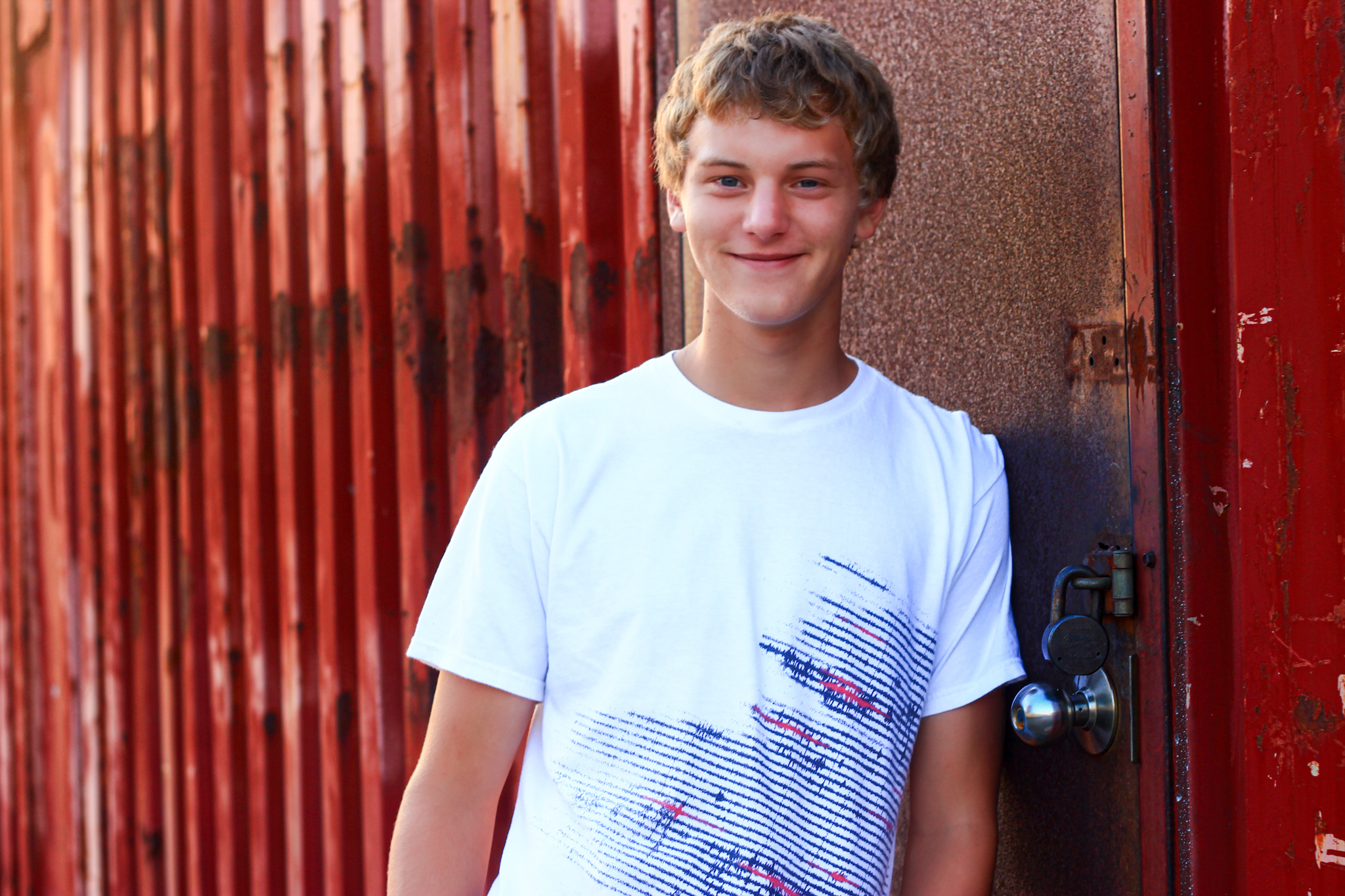 Young man leans against red metal building and smiles for a senior photo