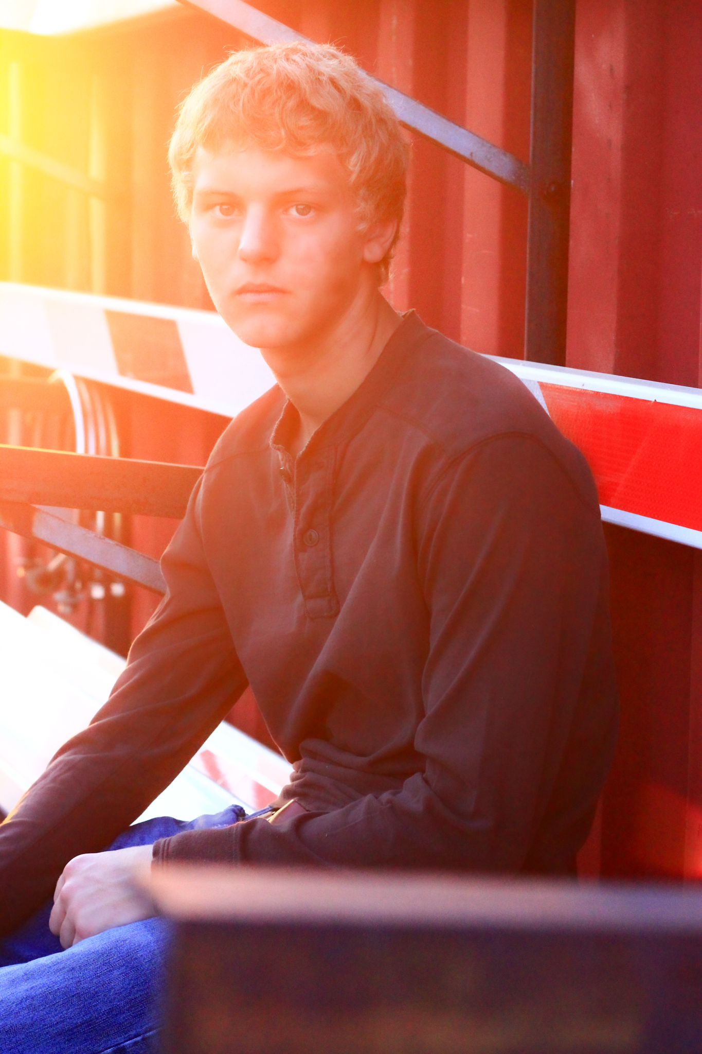 Young man sits against red metal building and arms of discarded railroad crossing during sunset for a senior photo