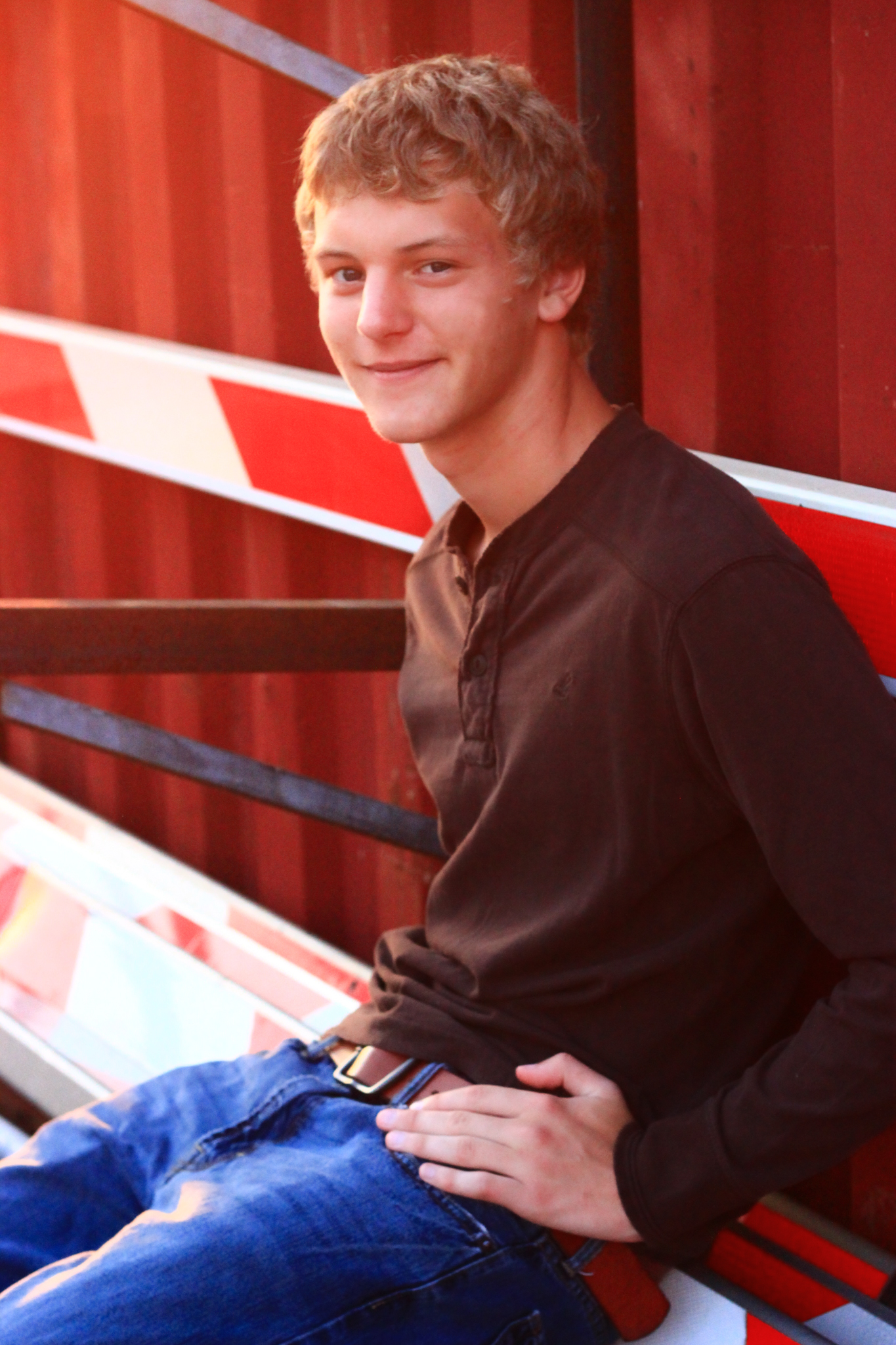 Young man sits against red metal building and arms of discarded railroad crossing during sunset smiling for a senior photo