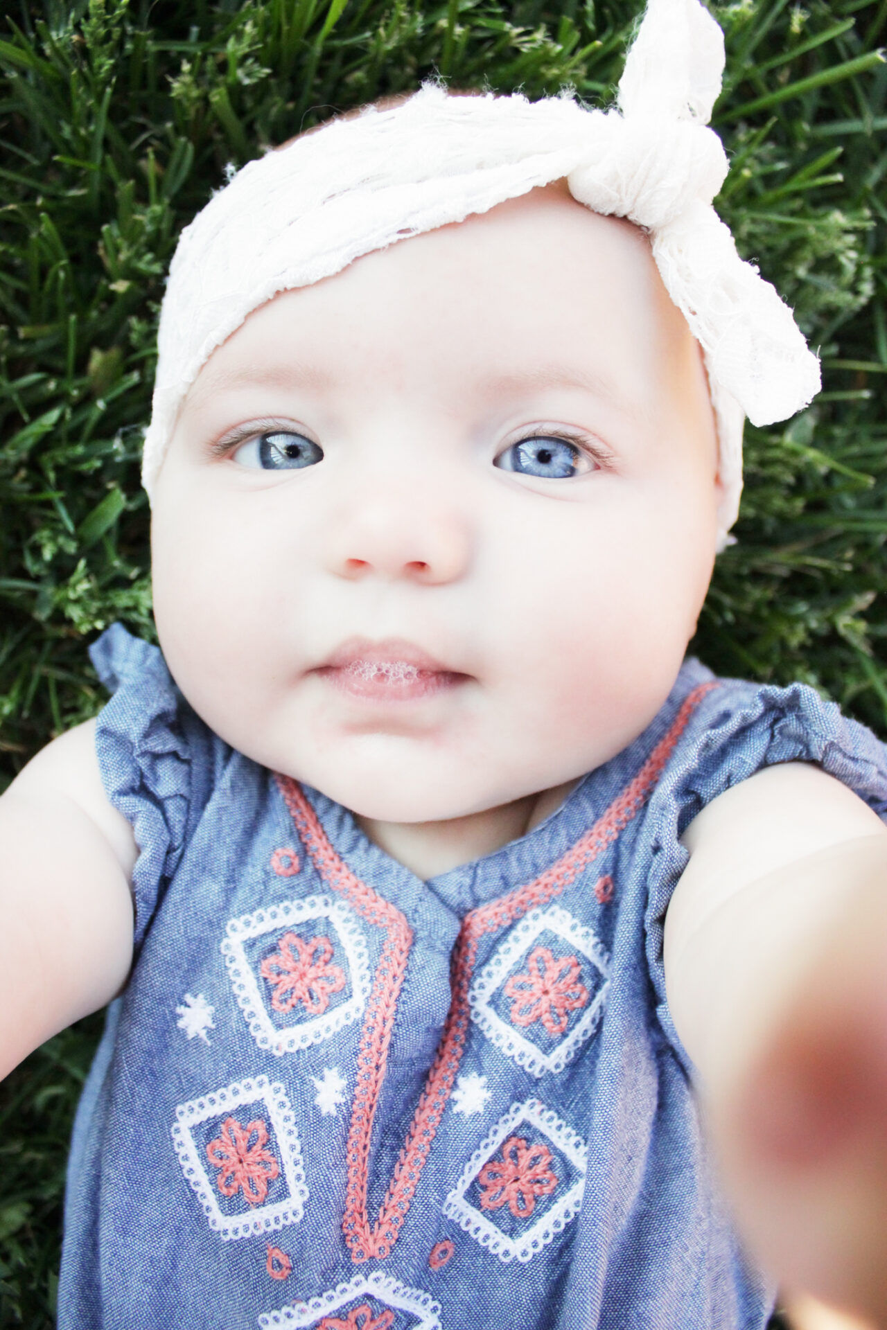 6 month old girl looking into camera while lying in the grass and blowing spit bubbles