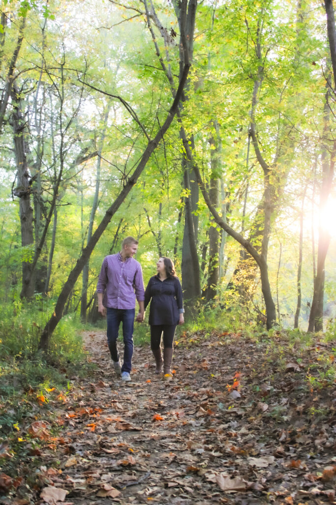 couple in maternity photo walk down a wooded pathway on leaves holding hands and looking at each other