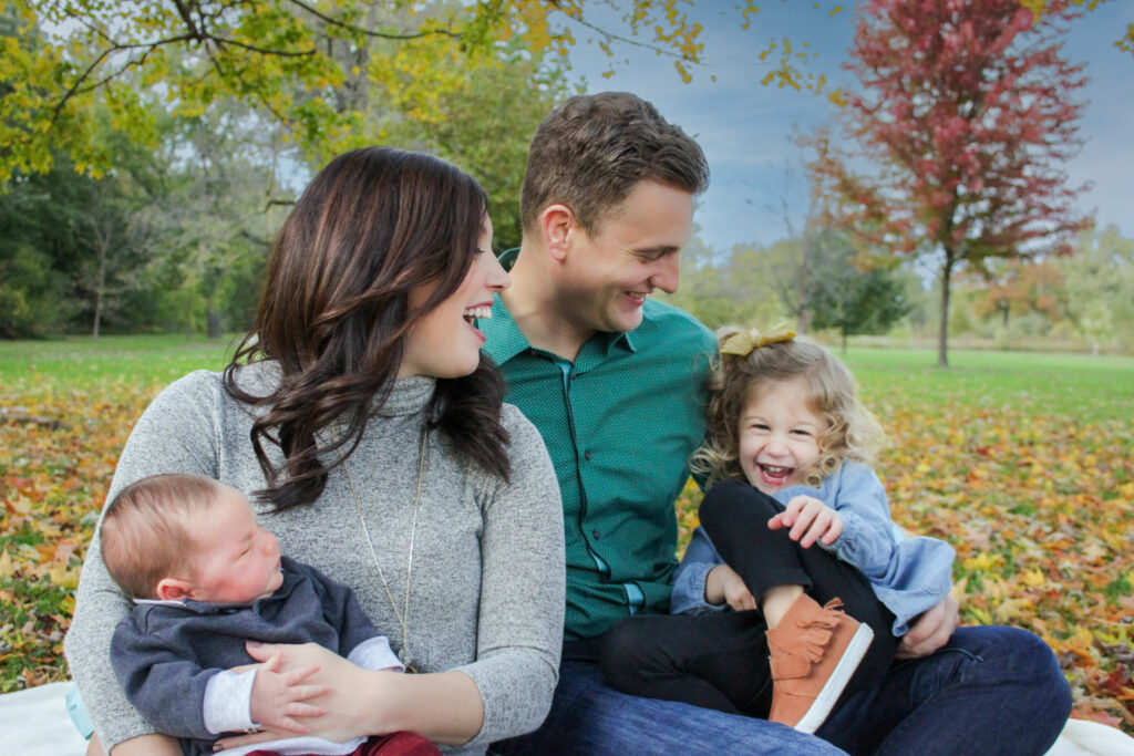 Family of four sitting on blanket in autumn leaves, father tickling little girl and mom holding baby boy