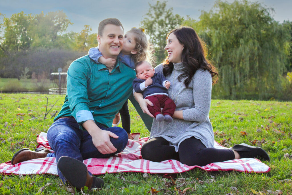 Family of four sitting on blanket in autumn leaves, daughter kissing father's cheek and mom holding baby boy