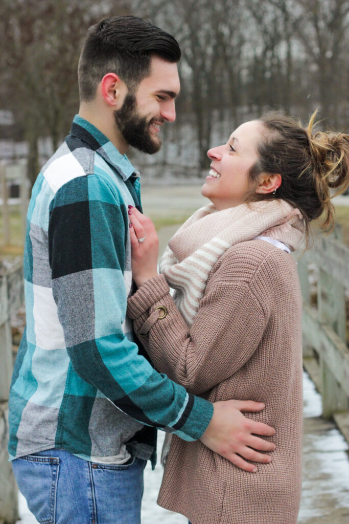 Young couple on wooden bridge in winter facing each other smiling for engagement photo