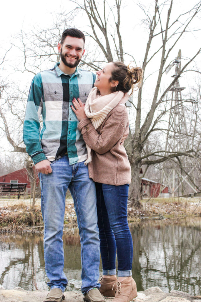 Young couple standing in front of pond in winter, girl has hand on boy's chest and is smiling looking at him, he is smiling at camera