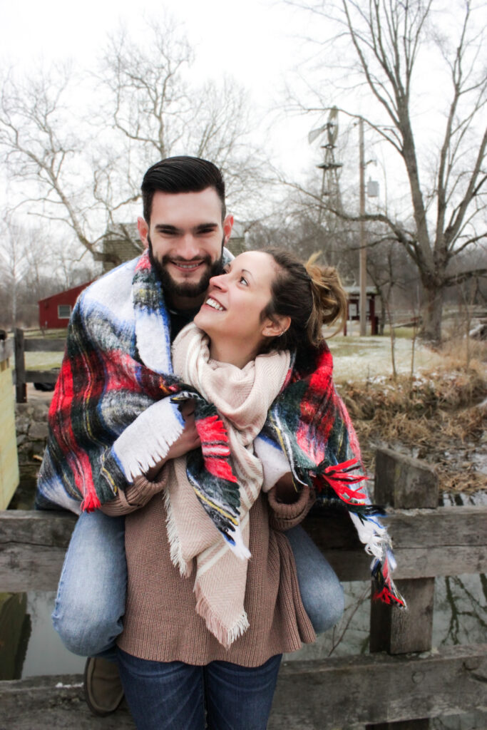 Young couple wrapped up in plaid blanket, boy is sitting on fence and smiling at camera and girl is in front of him smiling up at him