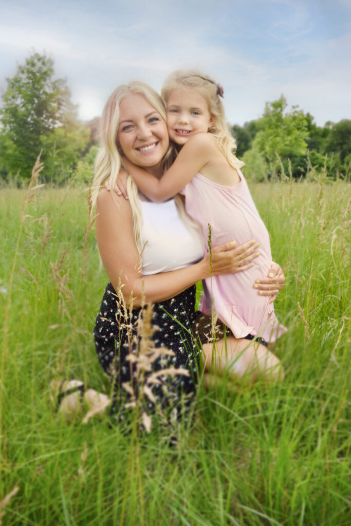 Young mother and daughter hug each other in grassy field and smile at camera during summer