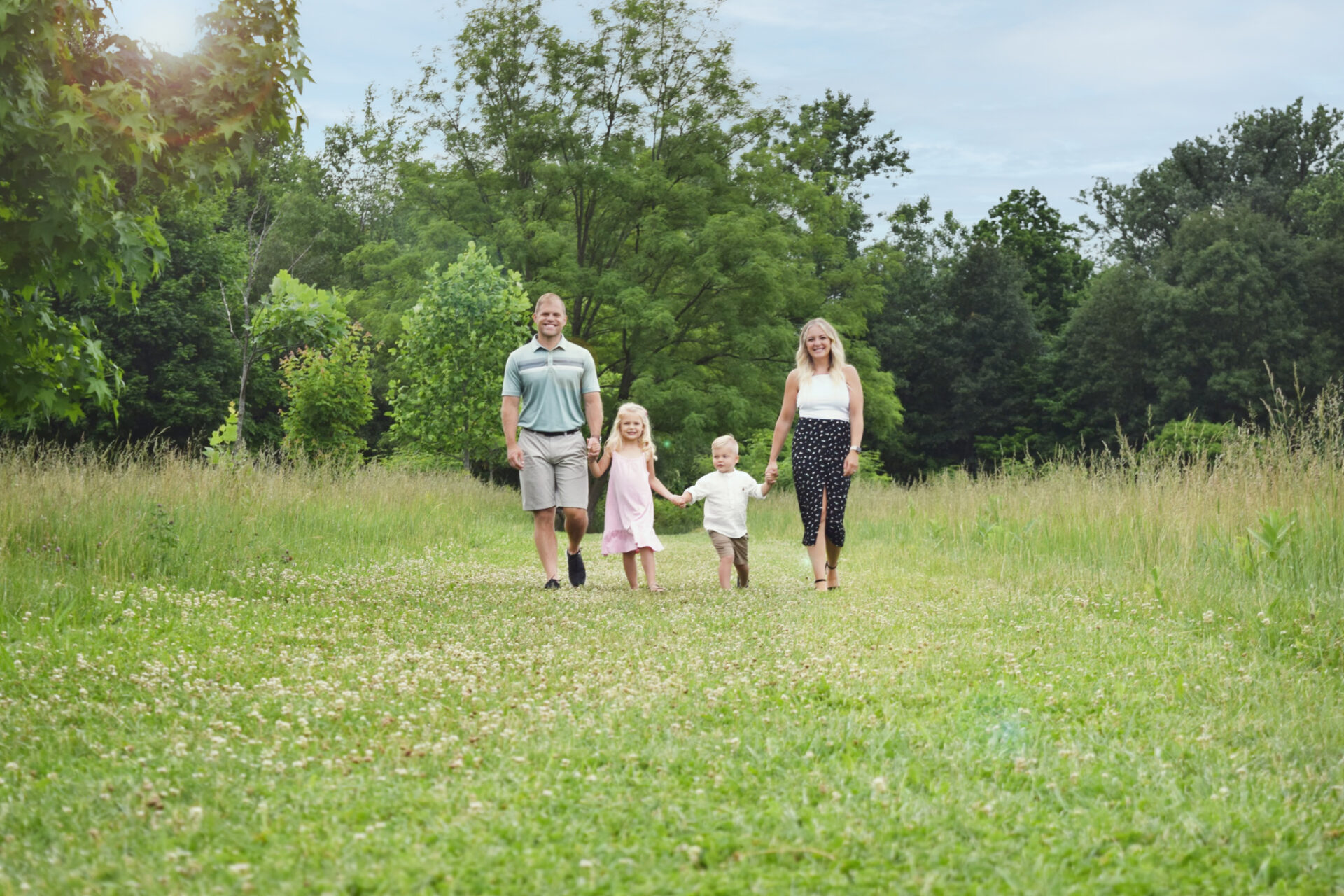 Young family of four holds hands and smiles at camera while walking on grassy path during summer