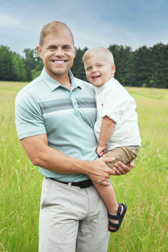 Young father holds young son in a grassy field during summer, both smile at camera