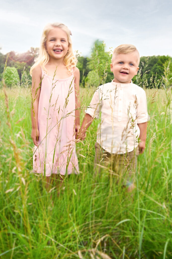 Young sister and brother hold hands and smiles at camera in grassy field during summer