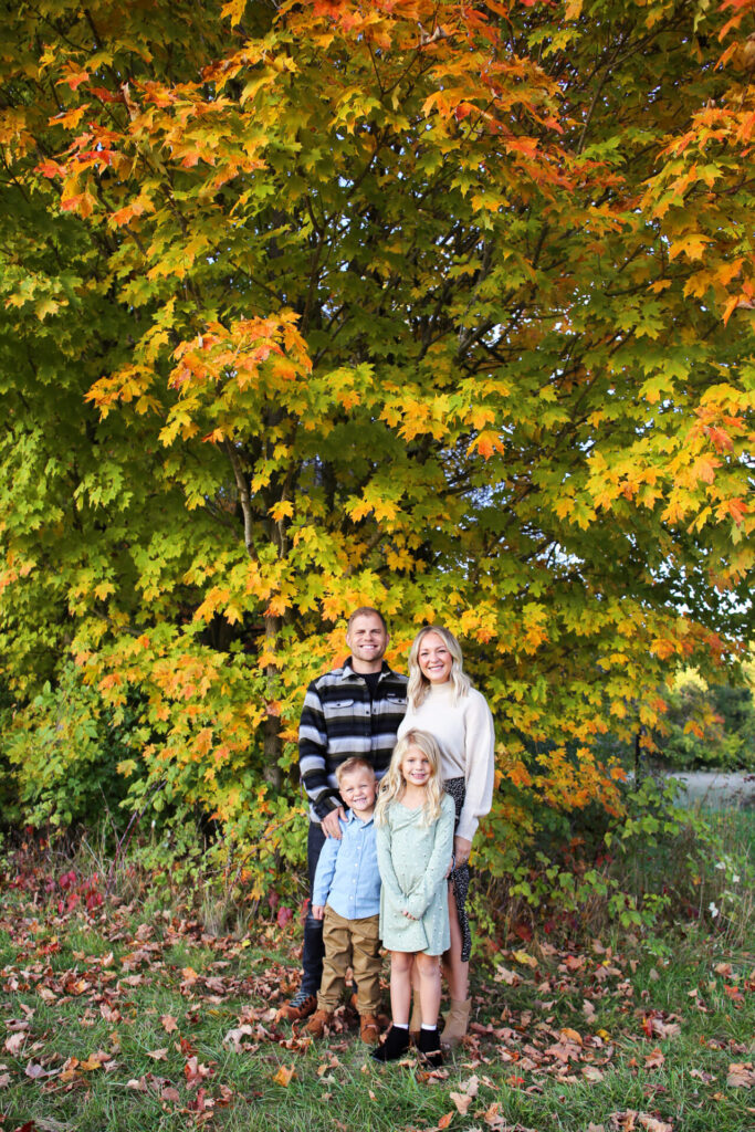 Young family of four stands together, smiling at camera, in front of tree in autumn