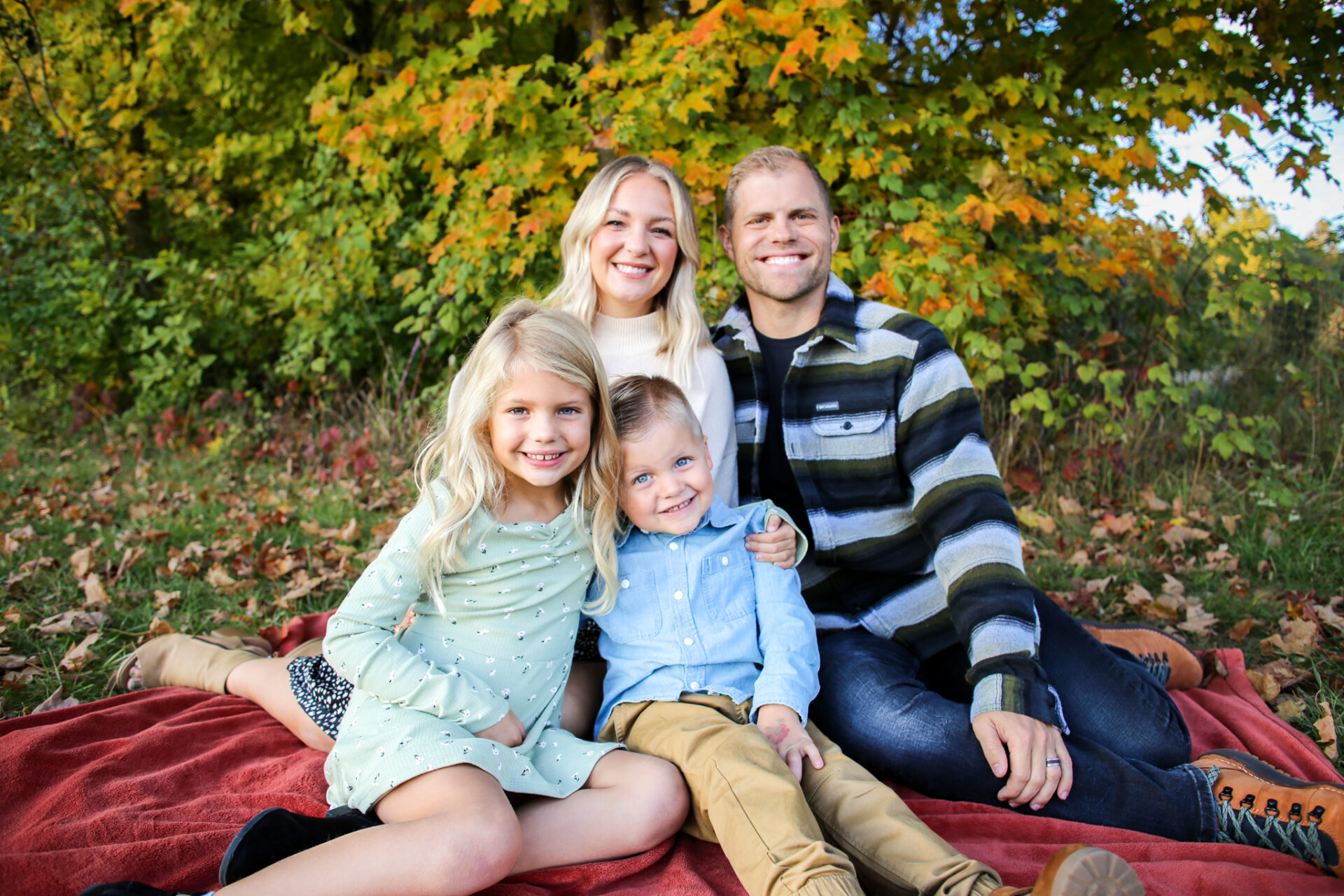 Young family of four sit on blanket together, smiling at camera, in front of tree in autumn
