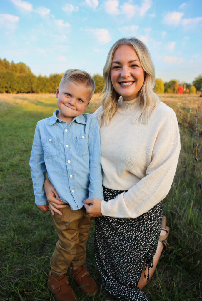 Young son stands with kneeling young mother, both smiling at camera, autumn