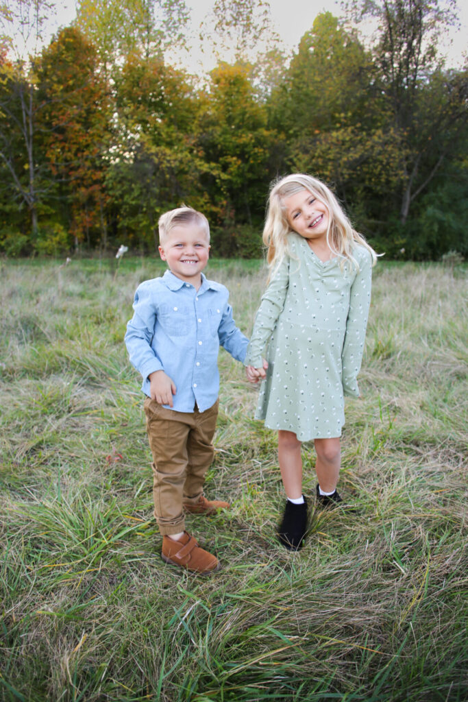 Young brother and sister hold hands and smile at camera in grassy field during autumn