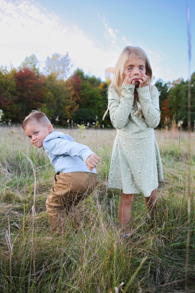 Young brother sticks bottom out at camera and young sister pulls eyes down for silly picture in a grassy field during autumn