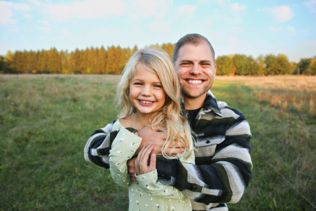 Young father wraps arms around young daughter, both smiling at camera during autumn