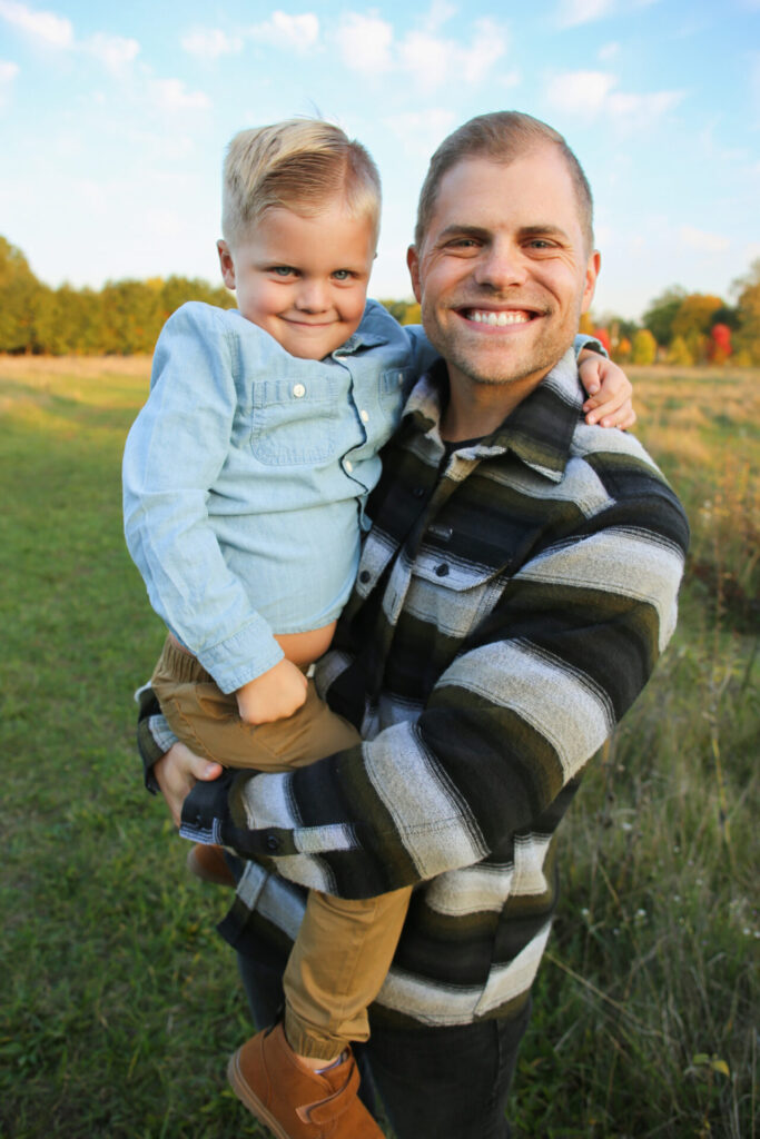 Young father holds young son and smiles at camera, boy looking mischievous