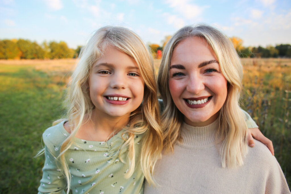 Young mother and daughter have arms around each other and smile at camera on a grassy path in autumn