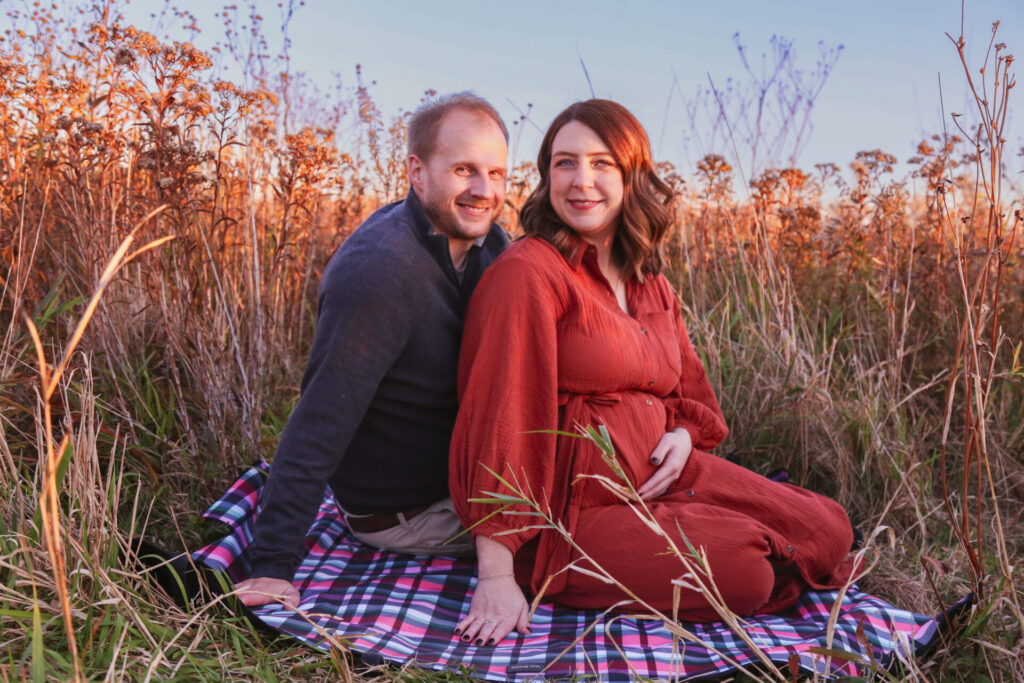 pregnant couple sit on blanket in tall grass and smile at camera